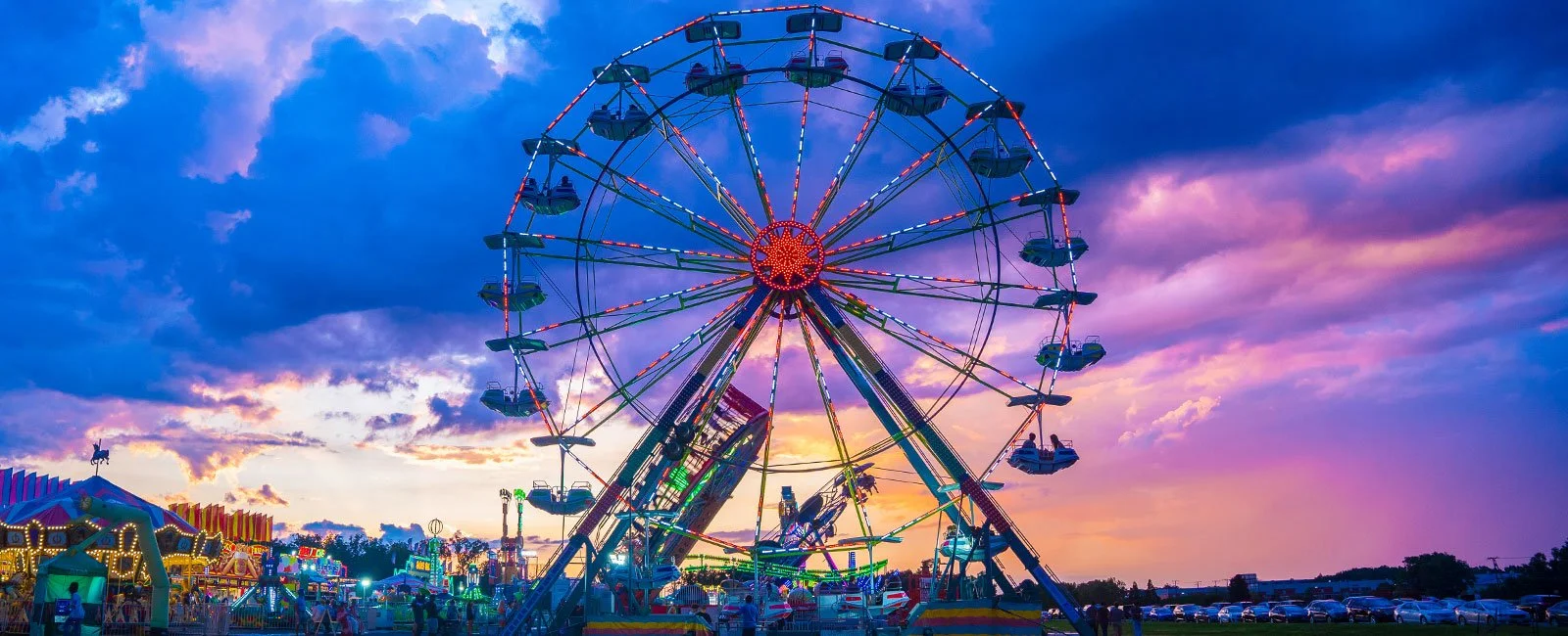 A brightly lit Ferris wheel and carnival midway beneath a colorful evening sky.